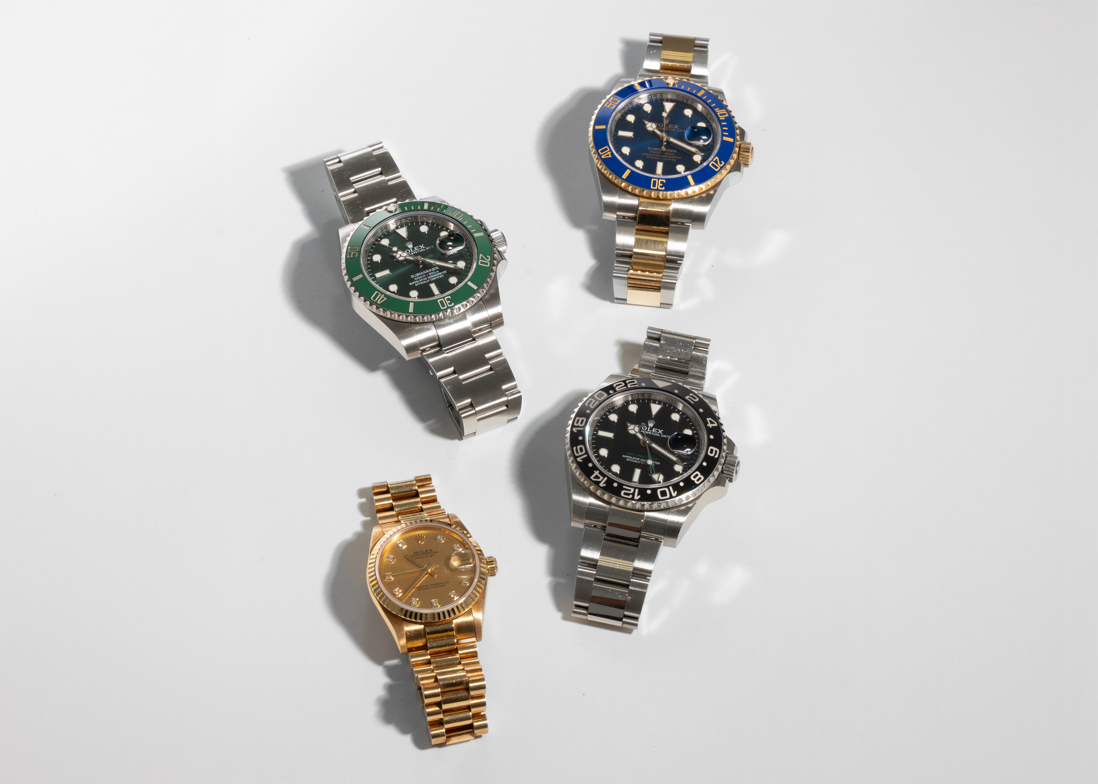 Four Rolex watches with different coloured faces and band materials on a light gray background